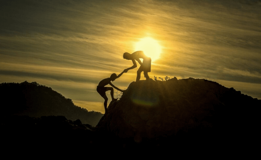 The image shows two people hiking a mountain and one of them is at the top helping the other person and giving them a boost during their final steps.