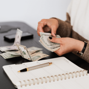 The image shows a woman's hands counting money and separating bills on the table in front of her. Also on the table are a notepad and pen. It's assumed that the woman is trying to figure out how much she'll have to pay out-of-pocket for counseling services.