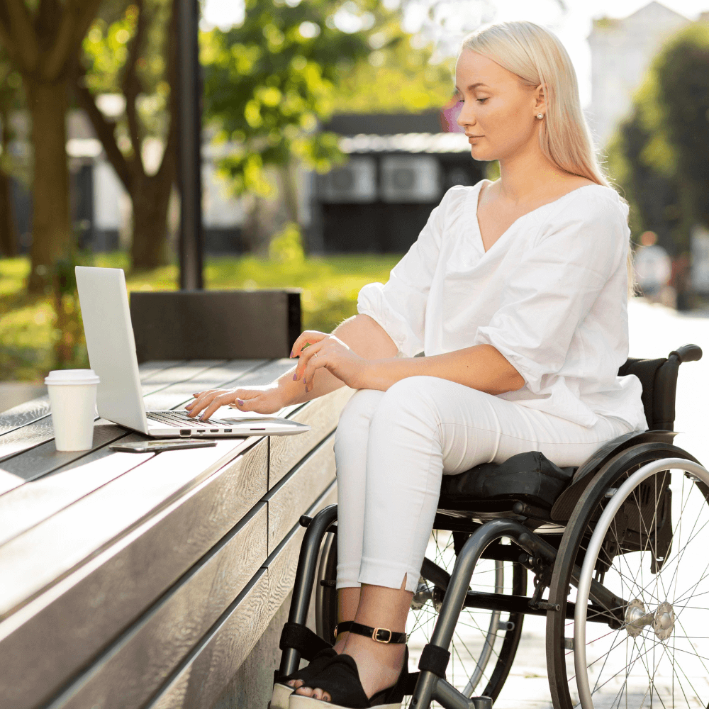 The image shows a woman in a wheelchair sitting outside and her computer is on a small bench in front of her. She's wearing all white and it looks to be sunny. It's assumed that the woman is on an online counseling session for suicide prevention therapy.
