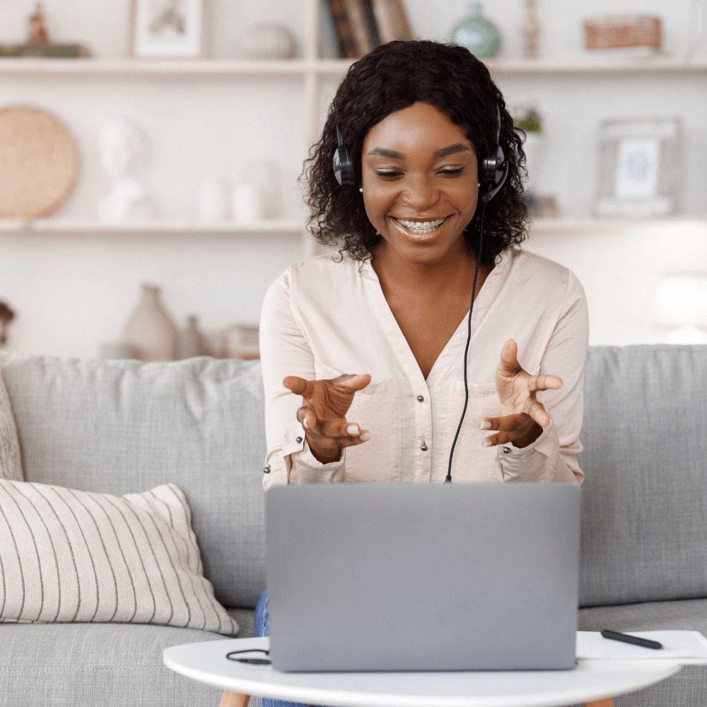 The image shows a Black woman sitting on a grey couch and smiling while looking at her computer. She has headphones on and it's assumed that she's undergoing therapy for suicide prevention.