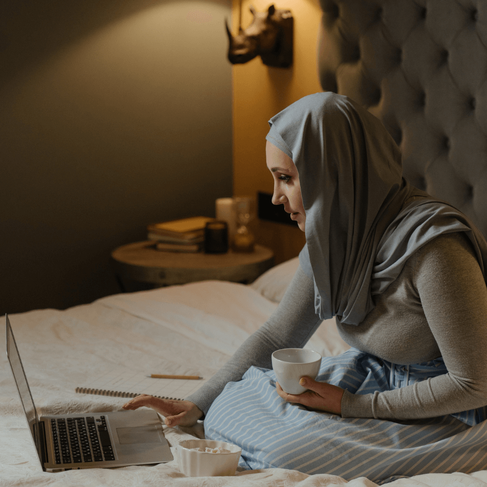 The image shows a woman in a hijab sitting on her bed and looking at her computer. It's assumed that she's in the middle of a virtual counseling session.