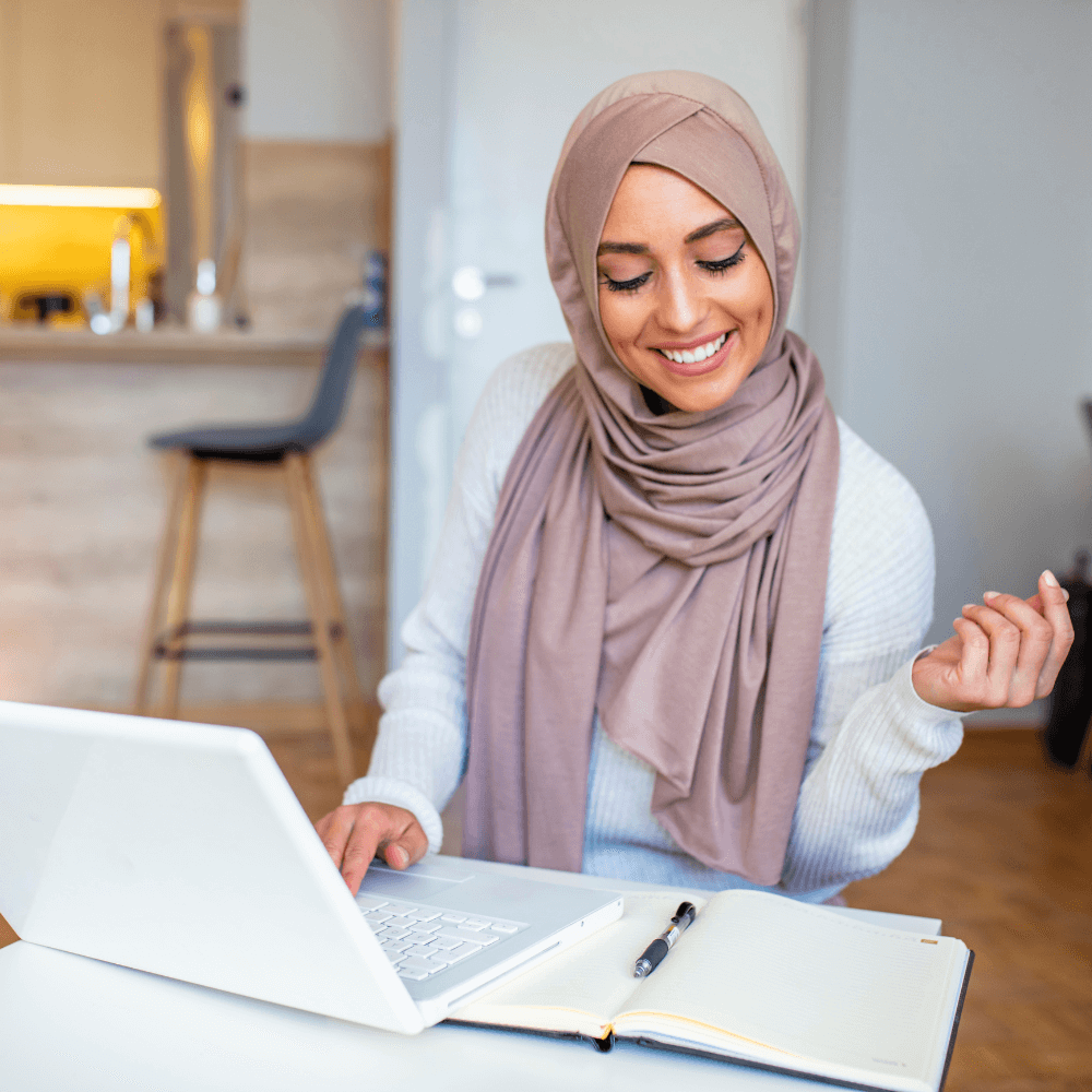 The image shows a woman wearing a hijab smiling while browsing her computer. In front of her on the table is also a notebook and pen.